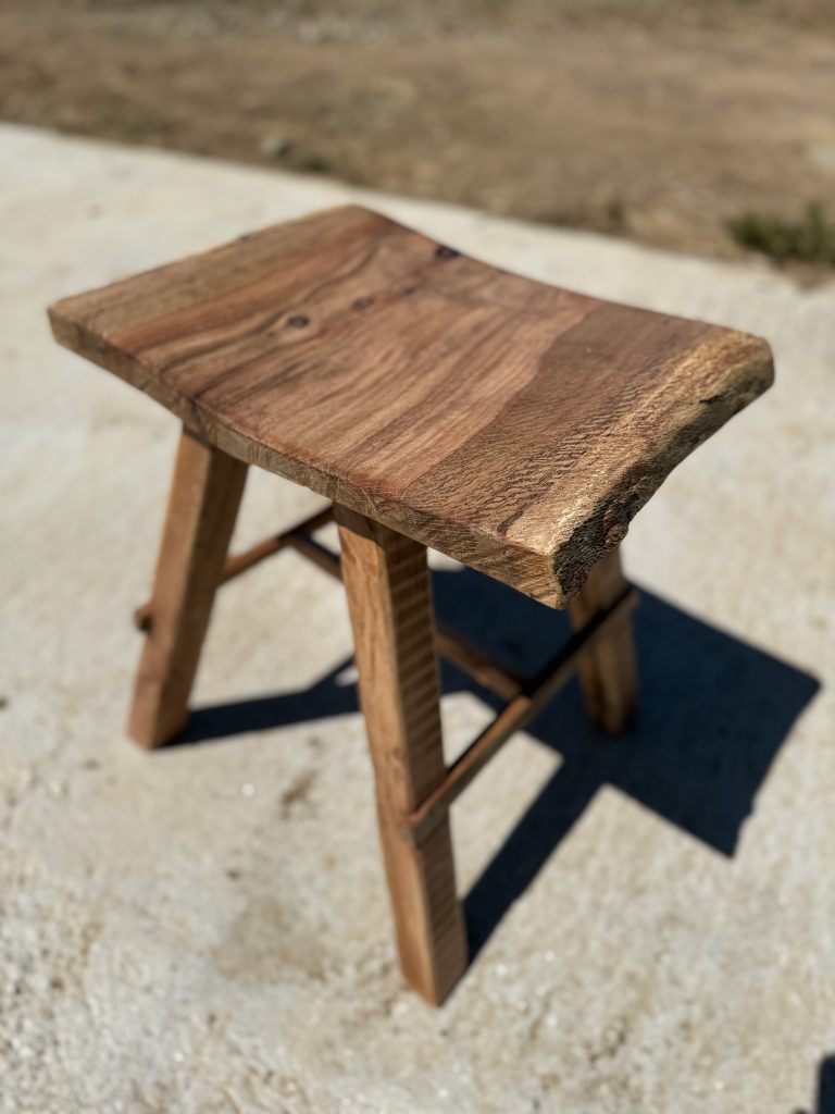 Taburete de madera con asiento rectangular de borde natural y cuatro patas.